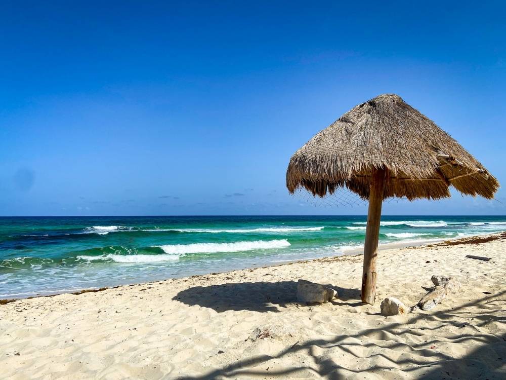 A traditional beach shelter in Cozumel during a Yucatan itinerary made with a straw roof and a wooden pole on sand with blue-green waves in the background