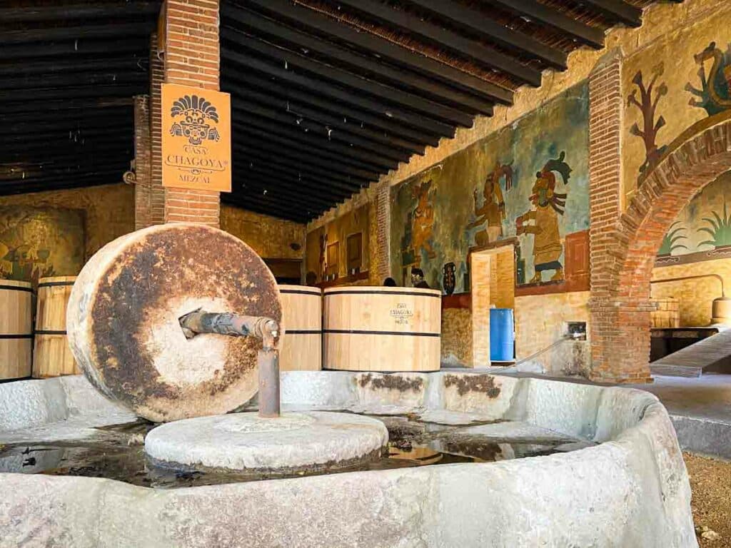 Upright stone wheel attached to a round platform for grinding. In the background are wooden barrels for mezcal and a wall with colorful Mayan images.