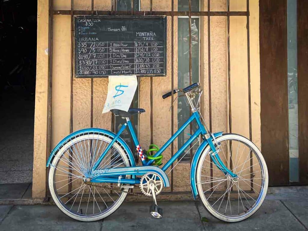 Blue metal two-wheeled bicycle leaning on a wall with a sign above it listing bike rental prices in Spanish