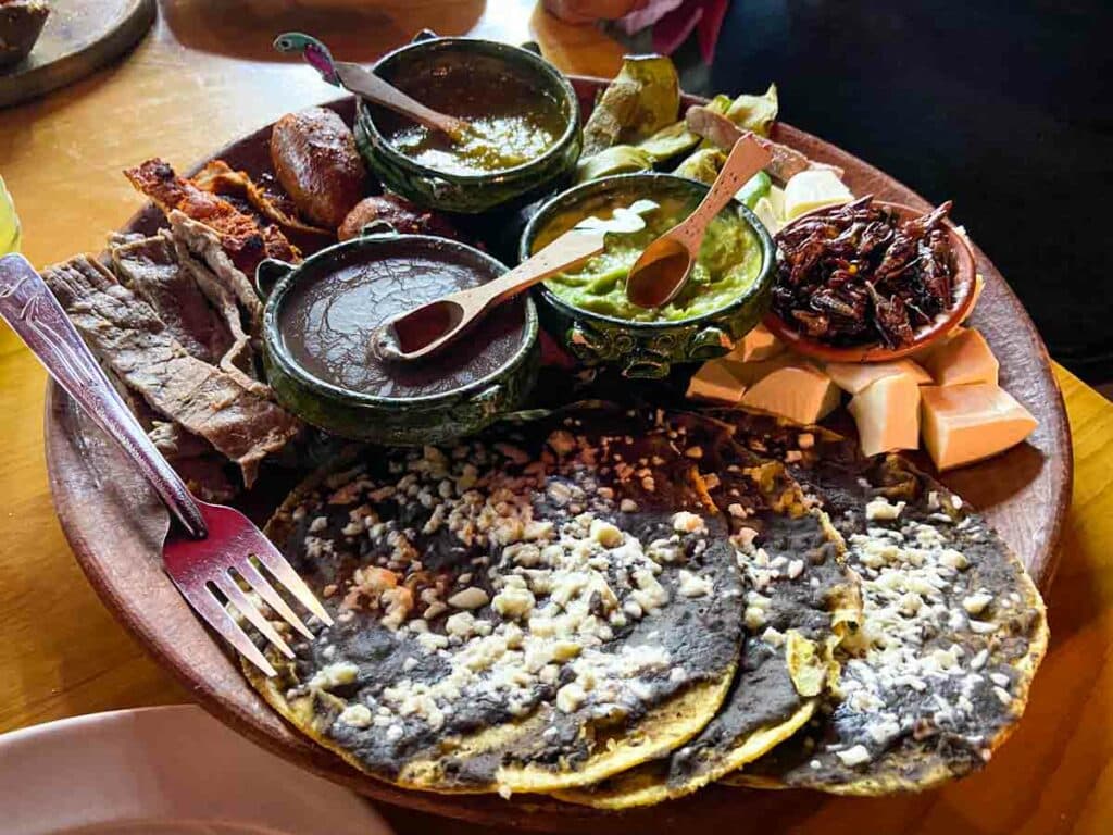 Ceramic platter on top of a wooden table covered with three thick corn tortillas topped with bean paste and cheese, three small bowls filled with salsa and wooden spoons, a small bowl of fried crickets on top of chopped jicama, roasted green vegetables, a variety of cooked meat strips, and a metal fork.
